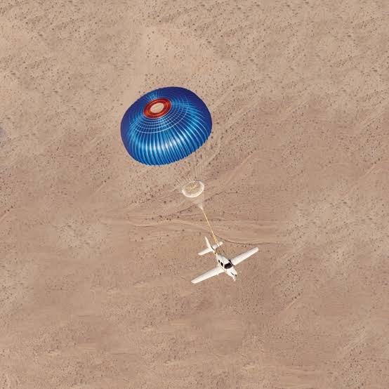 Aerial view of a Cirrus descending under its blue CAPS parachute over open desert